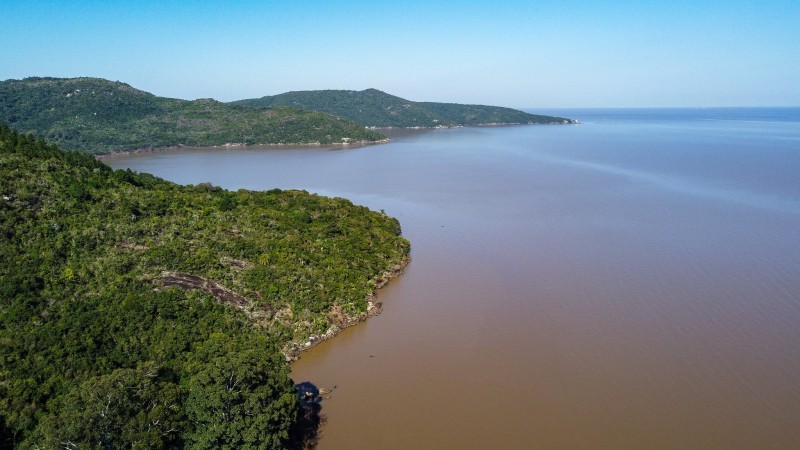 vista do morro do lago Guaíba - à esquerda trecho de terras coberto por árvore e à direito as águas do guaíba em cores mescladas em laranja e azul