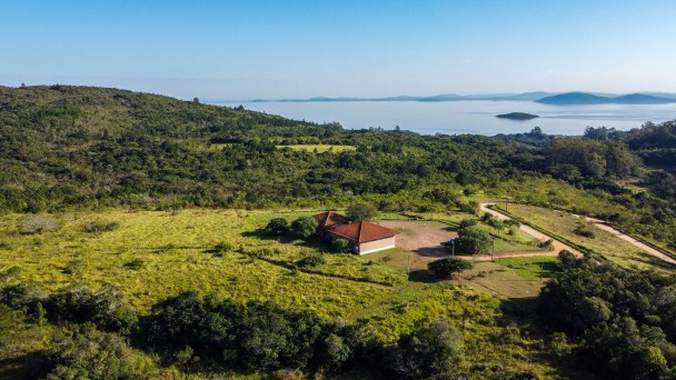 vista aérea de uma casa antiga de alvenaria ao centro, rodeada de vegetação e morro. Ao fundo, o lago guaíba.