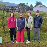 Três alunos de escolas públicas estão em pé, olhando para a câmera, ao lado de uma muda de árvore recém plantada. Ao centro, a diretora da escola.