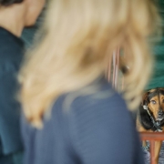 ao fundo da foto, um cachorro com o rosto caramelo e corpo com pelagem preta olha para frente. em primeiro plano, desfocado, um casal observa o cachorrinho.