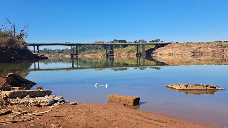 imagem mostra o rio Taquari Antas com um banco de detritos no centro e nas laterais. ao fundo uma ponte com veículos passando em cima. 