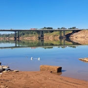 imagem mostra o rio Taquari Antas com um banco de detritos no centro e nas laterais. ao fundo uma ponte com veículos passando em cima. 