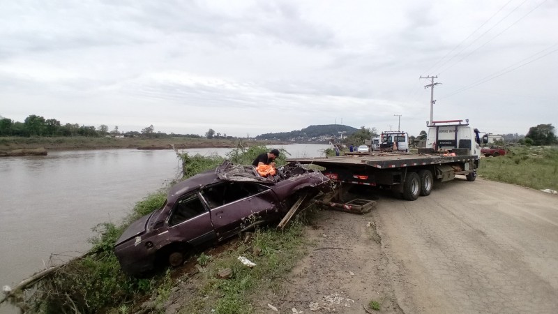 a imagem mostra o rio taquari à esquerda. ao centro, um carro da cor roxa que foi retirado do leito do rio. puxando o veículo, um caminhão rebocador sobre a estrada.