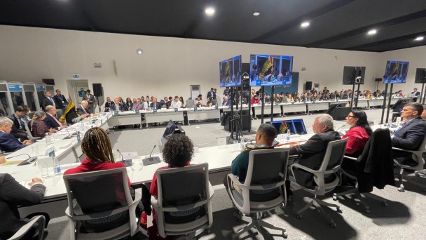 ao redor de uma mesa quadrada da cor branca autoridades brasileiras e da sociedade civil estão sentadas em volta, olhando para o vice-presidente da república, geraldo alckmin, que fala ao lado de ministros. ao centro, televisões transmitem a reunião.