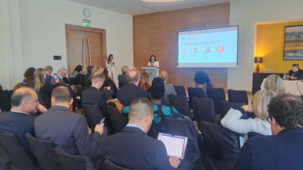 na imagem, a secretária Marjorie Kauffmann está ao fundo falando para uma plateia sentada. o evento aconteceu em uma sala, em um hotel de Baku, Azerbaijão