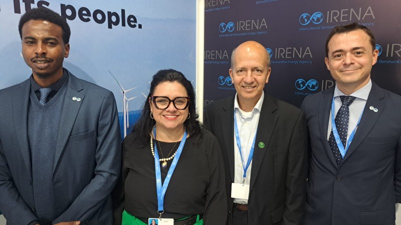 na imagem, quatro pessoas aparecem sorrindo para a câmera. ao centro, a secretária marjorie e o presidente Renato. nas extremidades, os representantes da Agência Internacional de Energia Renovável. Ao fundo, um painel com as marcas da instituição.