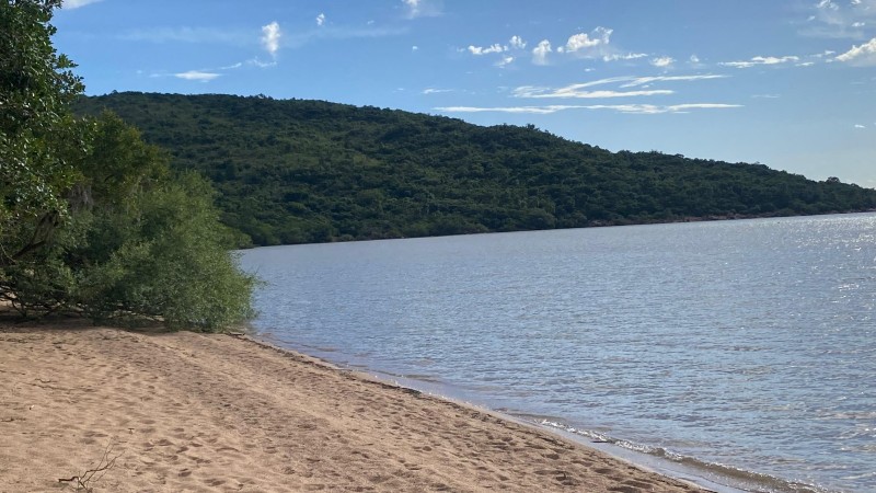 A imagem mostra a Praia das Pombas. à direita da imagem, a água calma do rio e à esquerda diversas pegadas na areia. o céu está azul com poucas núvens, ao fundo aparece um morro cheio de árvores verdes. 