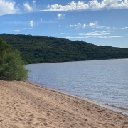 A imagem mostra a Praia das Pombas. à direita da imagem, a água calma do rio e à esquerda diversas pegadas na areia. o céu está azul com poucas núvens, ao fundo aparece um morro cheio de árvores verdes. 