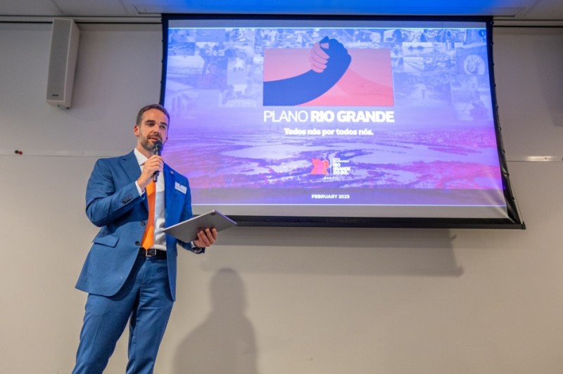 Governador Eduardo Leite, no lado esquerdo de um telão, com uma apresentação sobre o Plano Rio Grande. O governador veste um terno azul, camisa branca e uma gravata laranja.