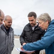 Grupo de quatro pessoas analisando um mapa ou projeto. Na esquerda, um homem de casaco preto, outro de casaco cinza, que é o secretário da Reconstrução Gaúcha, o governador Eduardo Leite, de preto, e uma senhora, mostrando o projeto, de cabelos brancos curtos e casaco azul, com capuz.