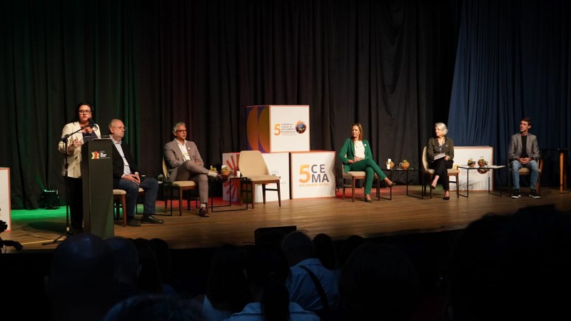 a imagem mostra o palco da associação médica do rio grande do sul. à esquerda, a secretária marjorie está em pé, em frente a um pulpito. sentados, autoridades que participam da conferência estadual do meio ambiente. ao centro ao fundo constam cubos com a marca da conferência.