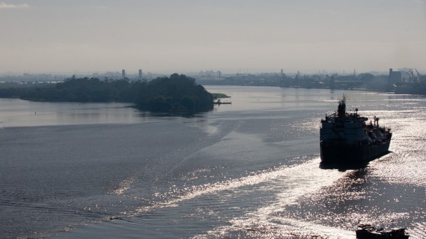 Embarcação de grande porte navega por um rio de águas calmas, com o reflexo do sol na superfície. À esquerda, uma pequena ilha coberta por vegetação. Ao fundo, estruturas industriais se destacam na linha do horizonte, parcialmente encobertas por neblina leve. O céu está claro, contribuindo para a visibilidade da paisagem fluvial.
