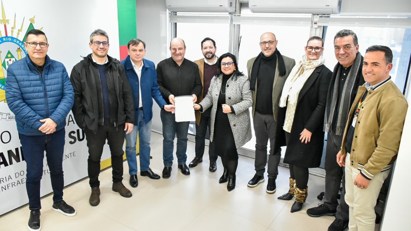 Grupo de 11 pessoas posa para foto em ambiente interno iluminado, em frente a uma parede com o brasão do Estado do Rio Grande do Sul e o texto “Governo do Estado do Rio Grande do Sul – Secretaria do Meio Ambiente e Infraestrutura”. Ao centro, duas pessoas seguram um documento com o memorando de entendimento assinado. o fundo, há janelas com persianas e dois aparelhos de ar-condicionado.
