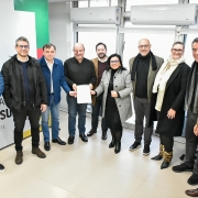 Grupo de 11 pessoas posa para foto em ambiente interno iluminado, em frente a uma parede com o brasão do Estado do Rio Grande do Sul e o texto “Governo do Estado do Rio Grande do Sul – Secretaria do Meio Ambiente e Infraestrutura”. Ao centro, duas pessoas seguram um documento com o memorando de entendimento assinado. o fundo, há janelas com persianas e dois aparelhos de ar-condicionado.