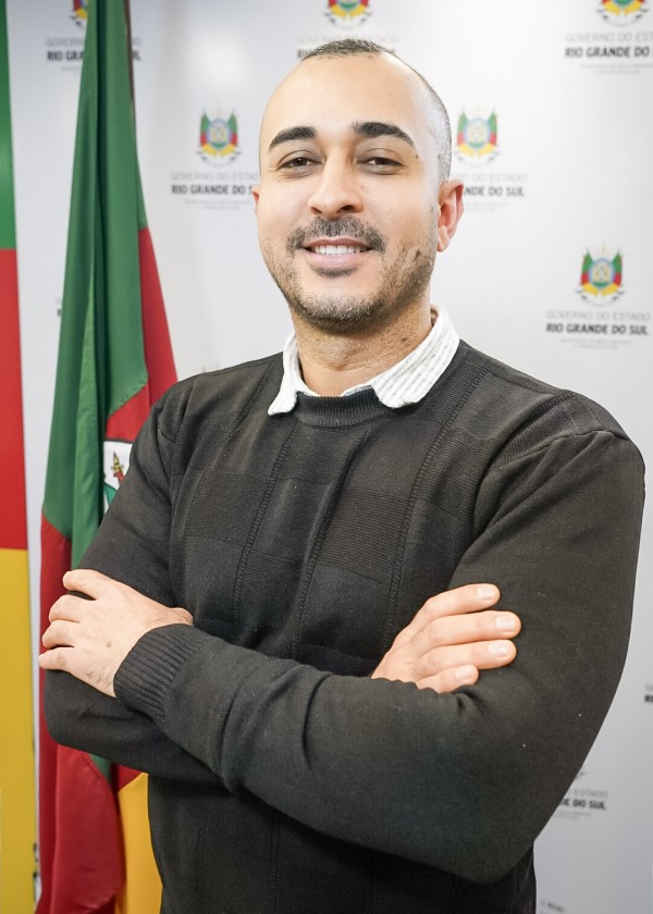 A imagem mostra um homem de pele clara, cabelo raspado e barba rala, sorrindo levemente com os braços cruzados. Ele está vestindo uma camisa clara com gola listrada sob um suéter preto. Ao fundo, há uma bandeira do Rio Grande do Sul parcialmente visível e um painel branco com o brasão e o texto “Governo do Estado do Rio Grande do Sul” repetido. A pose do homem transmite confiança e profissionalismo em um contexto institucional.