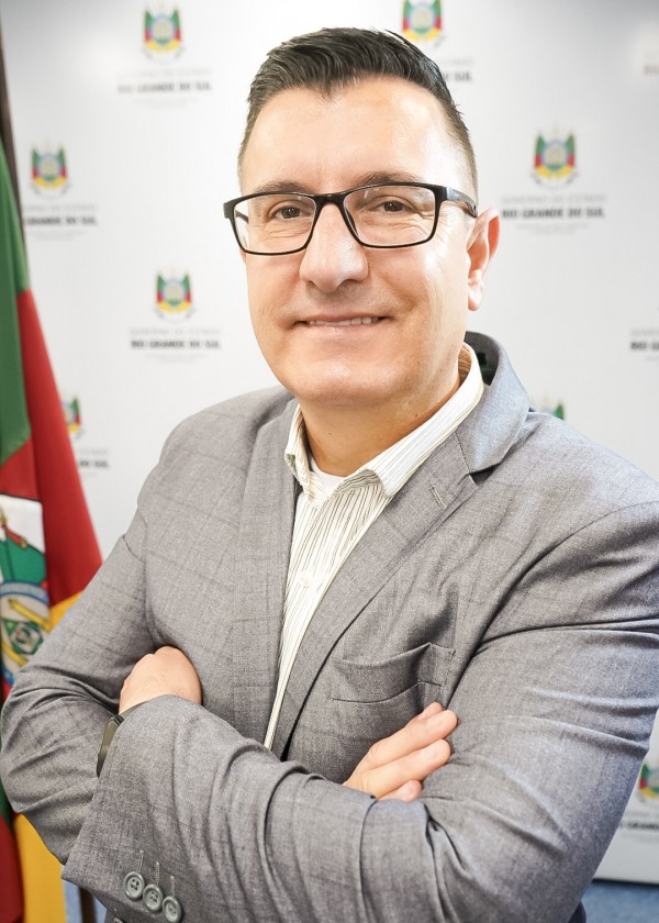 A imagem mostra um homem de pele clara, cabelos escuros e curtos penteados para o lado, usando óculos de armação preta. Ele está sorrindo levemente, com os braços cruzados, vestindo um paletó cinza claro sobre uma camisa social listrada em tons claros. Ao fundo, há uma bandeira do Rio Grande do Sul e um painel institucional branco com o brasão do estado e o texto “Governo do Estado do Rio Grande do Sul” repetido. A postura do homem transmite profissionalismo e confiança em um ambiente formal.