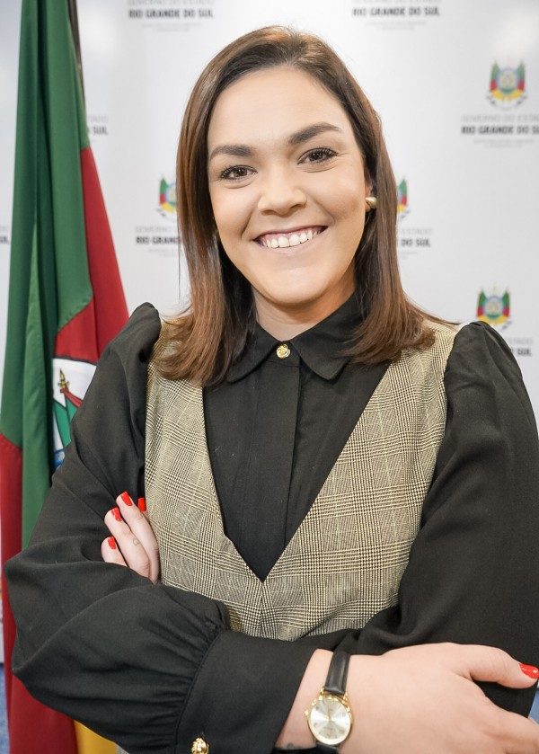 A imagem mostra uma mulher sorridente, de pele clara e cabelos castanhos lisos, na altura dos ombros. Ela está com os braços cruzados, usando uma blusa preta com mangas bufantes e botões dourados, e por cima, um colete xadrez em tons de bege e preto. Usa um relógio dourado com pulseira preta no pulso esquerdo e esmalte vermelho nas unhas. Ao fundo, há uma bandeira do Rio Grande do Sul e um painel institucional com o brasão e o texto “Governo do Estado do Rio Grande do Sul”. A imagem transmite uma postura segura e profissional em ambiente oficial.DSC05708 1