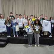 Grupo de 19 estudantes, meninos e meninas, posam sorrindo em um palco, segurando flores, sacolas e certificados. Alguns vestem camisetas brancas e outros roupas casuais. À esquerda, duas estudantes seguram uma faixa da Escola Municipal de Ensino Fundamental Nelly Dahne Logemann, com a frase “Educação: Somos Todos Responsáveis”. Ao fundo, cortina bege cobre toda a parede.
