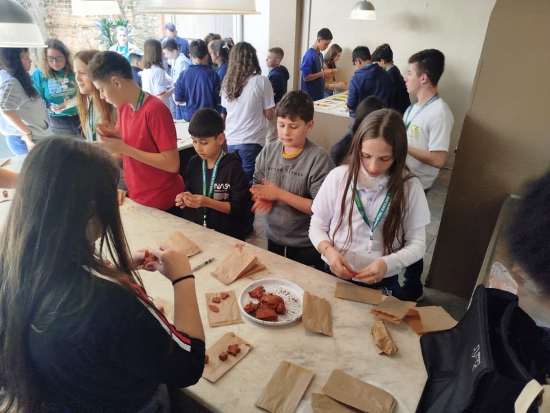 Grupo de crianças e adolescentes participa de uma atividade prática em um ambiente interno iluminado. Eles estão em pé ou sentados ao redor de mesas, manuseando pequenas porções de argila ou terra sobre folhas de papel pardo. Ao fundo, outros estudantes interagem e realizam atividades em outra mesa. O clima é de concentração e colaboração.
