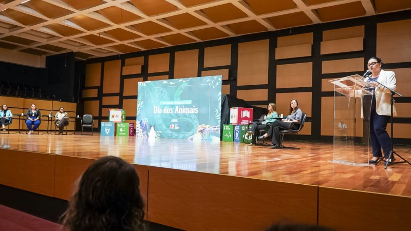 Palestrante fala ao microfone em audit&oacute;rio durante o 1&ordm; Encontro Estadual pelo Dia dos Animais. No palco, h&aacute; um grande painel com o nome do evento e outras participantes sentadas em cadeiras, compondo a mesa de abertura. O ambiente &eacute; iluminado e tem piso de madeira.