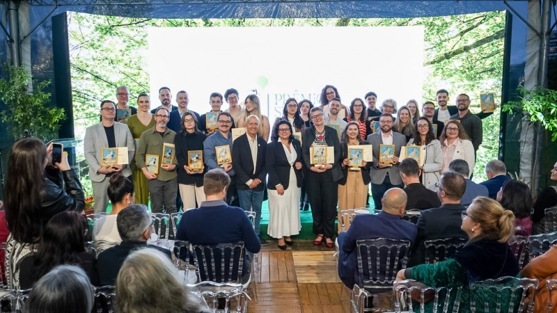 Grupo de pessoas em p&eacute; sobre um palco, segurando placas ou trof&eacute;us de premia&ccedil;&atilde;o. Ao fundo, h&aacute; um painel branco com logotipo e texto parcialmente vis&iacute;vel, indicando um evento formal de entrega de pr&ecirc;mios. Na frente do palco, v&aacute;rias pessoas est&atilde;o sentadas em cadeiras transparentes, assistindo &agrave; cerim&ocirc;nia. O ambiente &eacute; iluminado por luz natural, com &aacute;rvores vis&iacute;veis ao fundo, sugerindo que o evento ocorre em um espa&ccedil;o aberto ou com vista para a natureza.