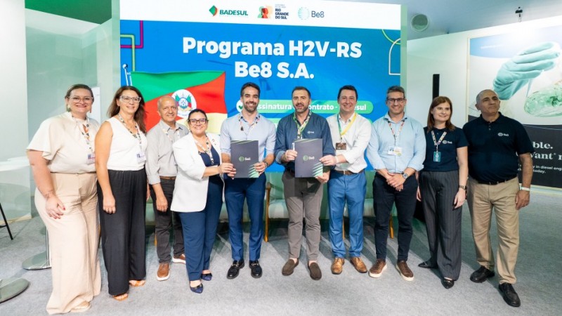 Oito pessoas estão alinhadas em frente a um painel azul com os logotipos da Badesul, Governo do Rio Grande do Sul e Be8. O texto principal diz “Programa H2V-RS Be8 S.A.” e abaixo “Assinatura de contrato Badesul”. Duas pessoas ao centro seguram pastas pretas com o logotipo da Be8, indicando a formalização do contrato. À esquerda do painel, aparece parcialmente a bandeira do Rio Grande do Sul. O ambiente é claro, com piso cinza e cadeiras ao fundo, típico de um evento corporativo.