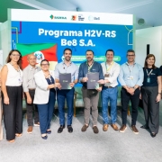 Oito pessoas estão alinhadas em frente a um painel azul com os logotipos da Badesul, Governo do Rio Grande do Sul e Be8. O texto principal diz “Programa H2V-RS Be8 S.A.” e abaixo “Assinatura de contrato Badesul”. Duas pessoas ao centro seguram pastas pretas com o logotipo da Be8, indicando a formalização do contrato. À esquerda do painel, aparece parcialmente a bandeira do Rio Grande do Sul. O ambiente é claro, com piso cinza e cadeiras ao fundo, típico de um evento corporativo.