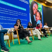 Seis pessoas estão sentadas em cadeiras de madeira com estofado claro, dispostas em linha sobre um tapete verde, participando de um painel de discussão. Ao fundo, há um grande telão azul com texto sobre política climática no Brasil, destacando temas como governança climática multissetorial e impactos da mudança do clima. À direita do telão, há uma imagem colorida de uma pessoa abraçando uma criança.