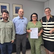 Quatro pessoas estão em pé dentro de um escritório, posicionadas lado a lado. A pessoa à esquerda veste uma camisa verde com o logotipo do IRGA e calça jeans. Ao lado, uma pessoa usa camisa social azul clara e calça cinza. A terceira pessoa veste um macacão verde e segura um documento impresso com o brasão do Estado do Rio Grande do Sul visível na parte superior. A quarta pessoa, à direita, está com um vestido preto e branco listrado horizontalmente, adornado com um colar de pérolas. Ao fundo, há duas fotografias emolduradas na parede e parte de um sofá preto.