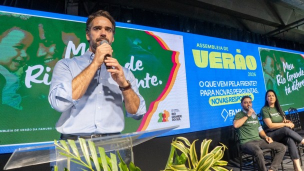 Homem segurando um microfone na m&atilde;o em primeiro plano, fundo colorido ao fundo e duas pessoas no canto direito da imagem.