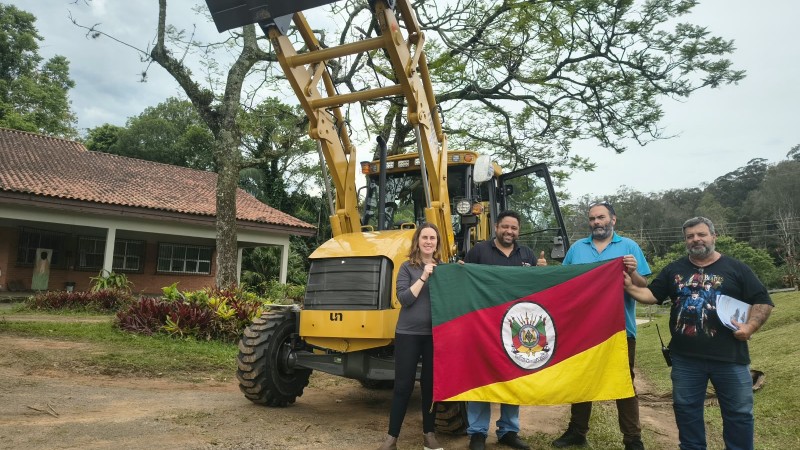 Uma mulher e tr&ecirc;s homens seguram a bandeira do Rio Grande do Sul em frente a uma retroescavadeira amarela. Ao fundo, &agrave; esquerda, uma casa t&eacute;rrea com telhado e tijolos &agrave; vista e, &agrave; direita, v&aacute;rias &aacute;rvores.
