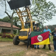 Uma mulher e tr&ecirc;s homens seguram a bandeira do Rio Grande do Sul em frente a uma retroescavadeira amarela. Ao fundo, &agrave; esquerda, uma casa t&eacute;rrea com telhado e tijolos &agrave; vista e, &agrave; direita, v&aacute;rias &aacute;rvores.